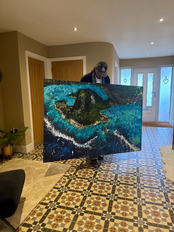 Ben holding a large commissioned canvas, an aerial island painting in deep teal