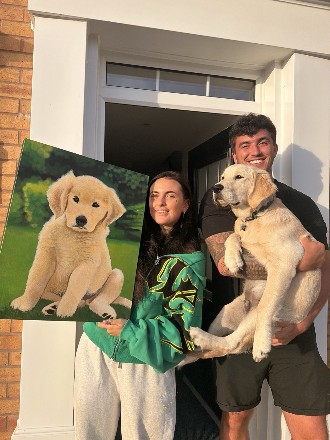 Niamh handing a client the commissioned portrait she painted of their golden retriever puppy, client and puppy beside the finished piece