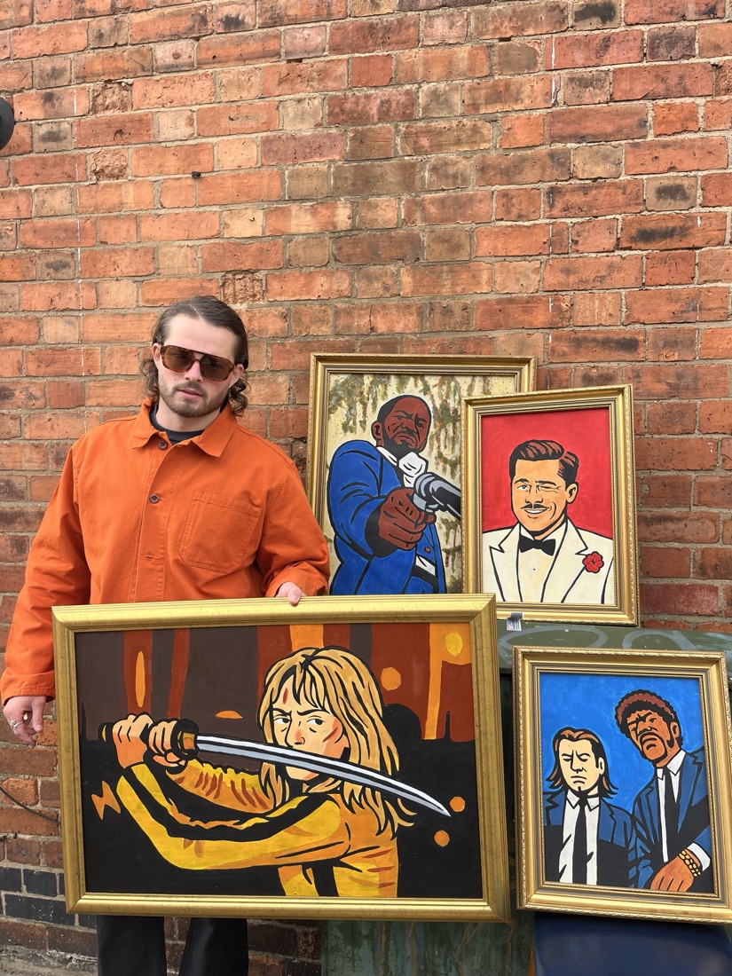 Ben beside his framed pop-culture portrait paintings on a brick wall
