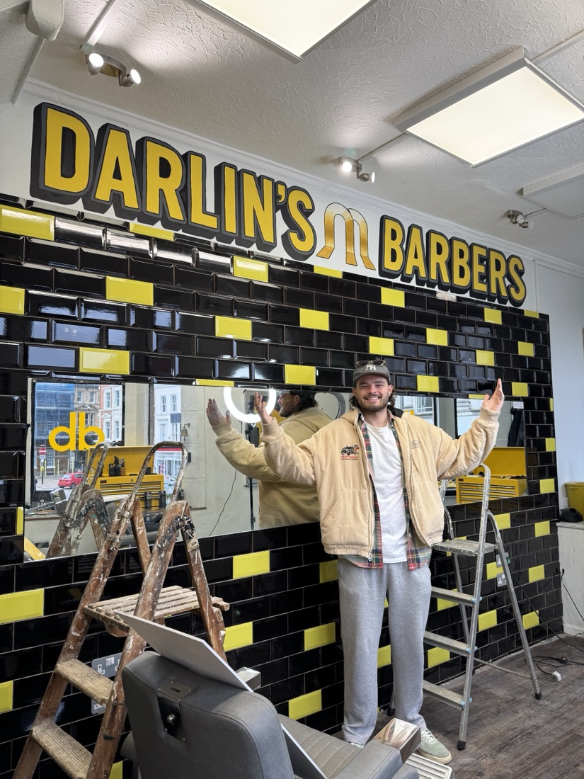 Ben in front of the Darlin's Barbers logo mural, black and yellow tiled wall